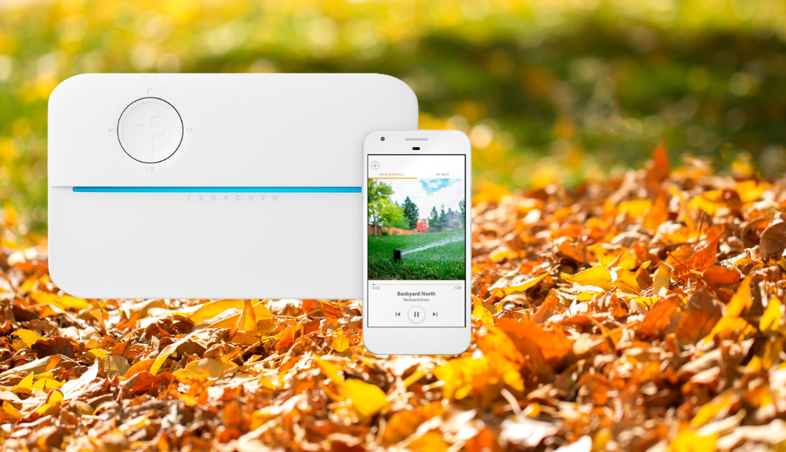 A picture of a white weather-based smart sprinkler timer and smartphone with a fall landscape of orange leaves in the background.